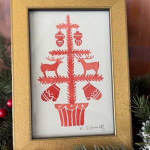 K&ouml;nnte beinhalten: Ein gerahmter Druck in einem goldfarbenen Rahmen. Der Druck zeigt ein rotes Weihnachtsbaumdesign mit Hirschen, Ornamenten und F&auml;ustlingen. Die Signatur des K&uuml;nstlers, "K.Schmitz", ist unten rechts sichtbar.