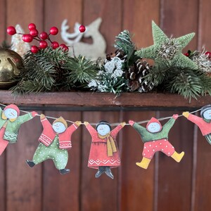 K&ouml;nnte beinhalten: Eine festliche Weihnachts-Girlande mit f&uuml;nf Papierfiguren in Winterkleidung, die sich an den H&auml;nden halten. Die Girlande ist unter einem Regal angebracht, das mit Tannenzweigen, roten Beeren, einem Stern und einer Glocke geschm&uuml;ckt ist.