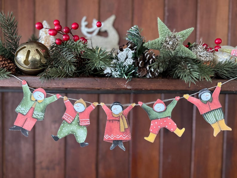 K&ouml;nnte beinhalten: Eine festliche Weihnachts-Girlande mit f&uuml;nf Papierfiguren in Winterkleidung, die sich an den H&auml;nden halten. Die Girlande ist unter einem Regal angebracht, das mit Tannenzweigen, roten Beeren, einem Stern und einer Glocke geschm&uuml;ckt ist.