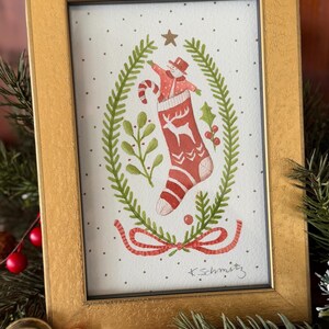 K&ouml;nnte beinhalten: Ein gerahmtes Aquarellgem&auml;lde mit einem roten Weihnachtsstrumpf mit Hirschmuster, einem Schneemann und einer Zuckerstange. Das Kunstwerk ist von einem gr&uuml;nen Kranz und einem goldenen Rahmen umgeben. Festliche Weihnachtsdekoration.