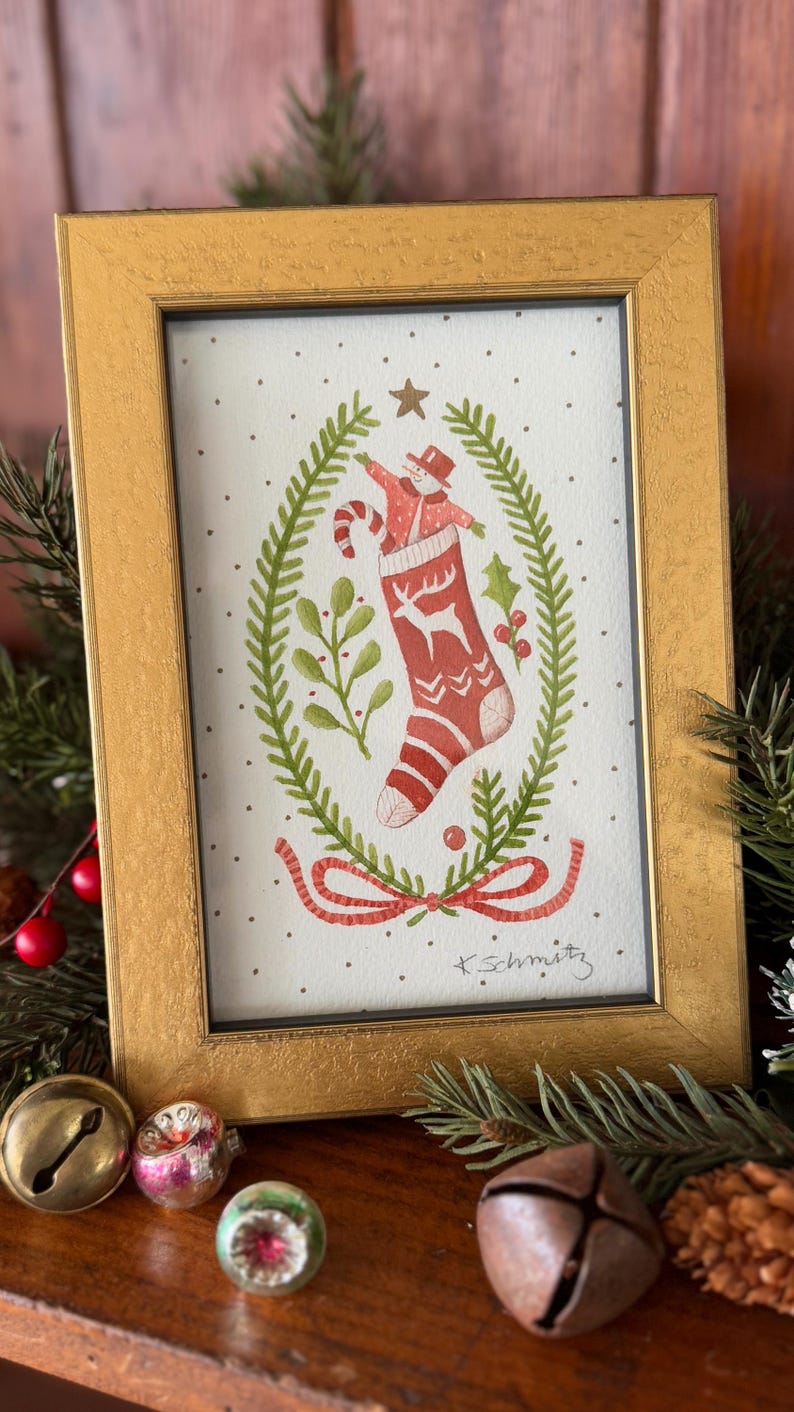 K&ouml;nnte beinhalten: Ein gerahmtes Aquarellgem&auml;lde mit einem roten Weihnachtsstrumpf mit Hirschmuster, einem Schneemann und einer Zuckerstange. Das Kunstwerk ist von einem gr&uuml;nen Kranz und einem goldenen Rahmen umgeben. Festliche Weihnachtsdekoration.