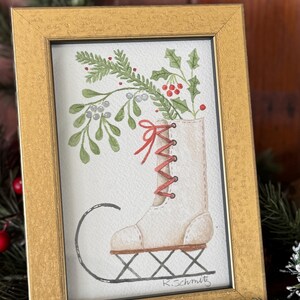 K&ouml;nnte beinhalten: Aquarellmalerei in einem goldenen Rahmen zeigt einen wei&szlig;en Schlittschuh, gef&uuml;llt mit Gr&uuml;n und roten Beeren. Der Schuh hat rote Schn&uuml;rsenkel und steht auf einem schwarzen L&auml;ufer. Die Signatur des K&uuml;nstlers, "K.Schmitz", ist sichtbar.