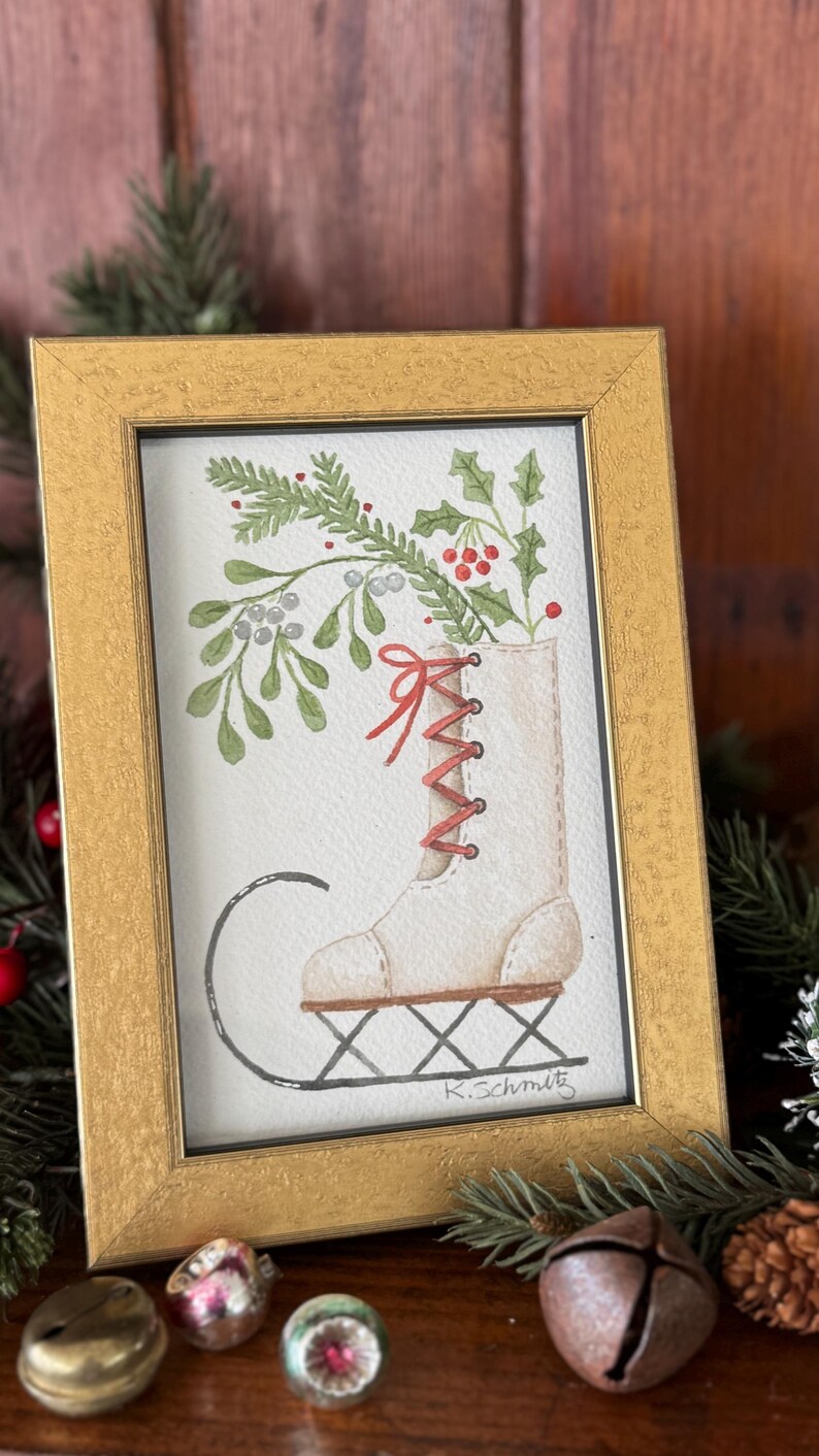 K&ouml;nnte beinhalten: Aquarellmalerei in einem goldenen Rahmen zeigt einen wei&szlig;en Schlittschuh, gef&uuml;llt mit Gr&uuml;n und roten Beeren. Der Schuh hat rote Schn&uuml;rsenkel und steht auf einem schwarzen L&auml;ufer. Die Signatur des K&uuml;nstlers, "K.Schmitz", ist sichtbar.