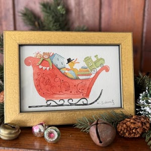 K&ouml;nnte beinhalten: Gerahmtes Aquarellgem&auml;lde eines roten Schlittens, gef&uuml;llt mit Geschenken und Spielzeug, darunter ein Teddyb&auml;r, ein Haus und ein Zug. Eine gelbe Ente mit Zylinder sitzt im Schlitten. Der Rahmen ist goldfarben.