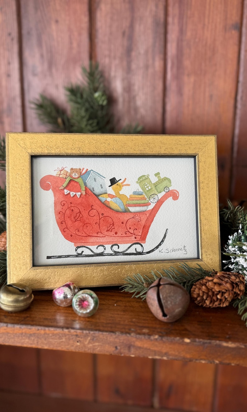 K&ouml;nnte beinhalten: Gerahmtes Aquarellgem&auml;lde eines roten Schlittens, gef&uuml;llt mit Geschenken und Spielzeug, darunter ein Teddyb&auml;r, ein Haus und ein Zug. Eine gelbe Ente mit Zylinder sitzt im Schlitten. Der Rahmen ist goldfarben.