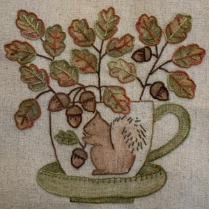 May include: Embroidered artwork of a squirrel sitting in a teacup with autumn leaves and acorns. The teacup is on a saucer.