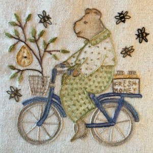 Może przedstawiać: Brązowy niedźwiedź jadący na niebieskim rowerze z koszykiem pełnym zielonych liści i żółtej gruszki. Rower ma z tyłu skrzynię z napisem "Świeży miód".