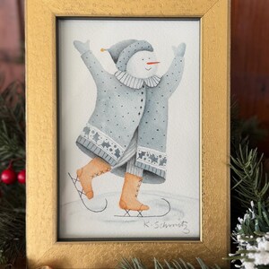 K&ouml;nnte beinhalten: Ein gerahmtes Aquarellgem&auml;lde eines Schneemanns beim Schlittschuhlaufen, der einen blauen Mantel und Hut sowie braune Schlittschuhe tr&auml;gt. Das Gem&auml;lde befindet sich in einem goldenen Rahmen, und die Signatur des K&uuml;nstlers befindet sich unten rechts. Weihnachtsdekorationen umgeben den Rahmen.