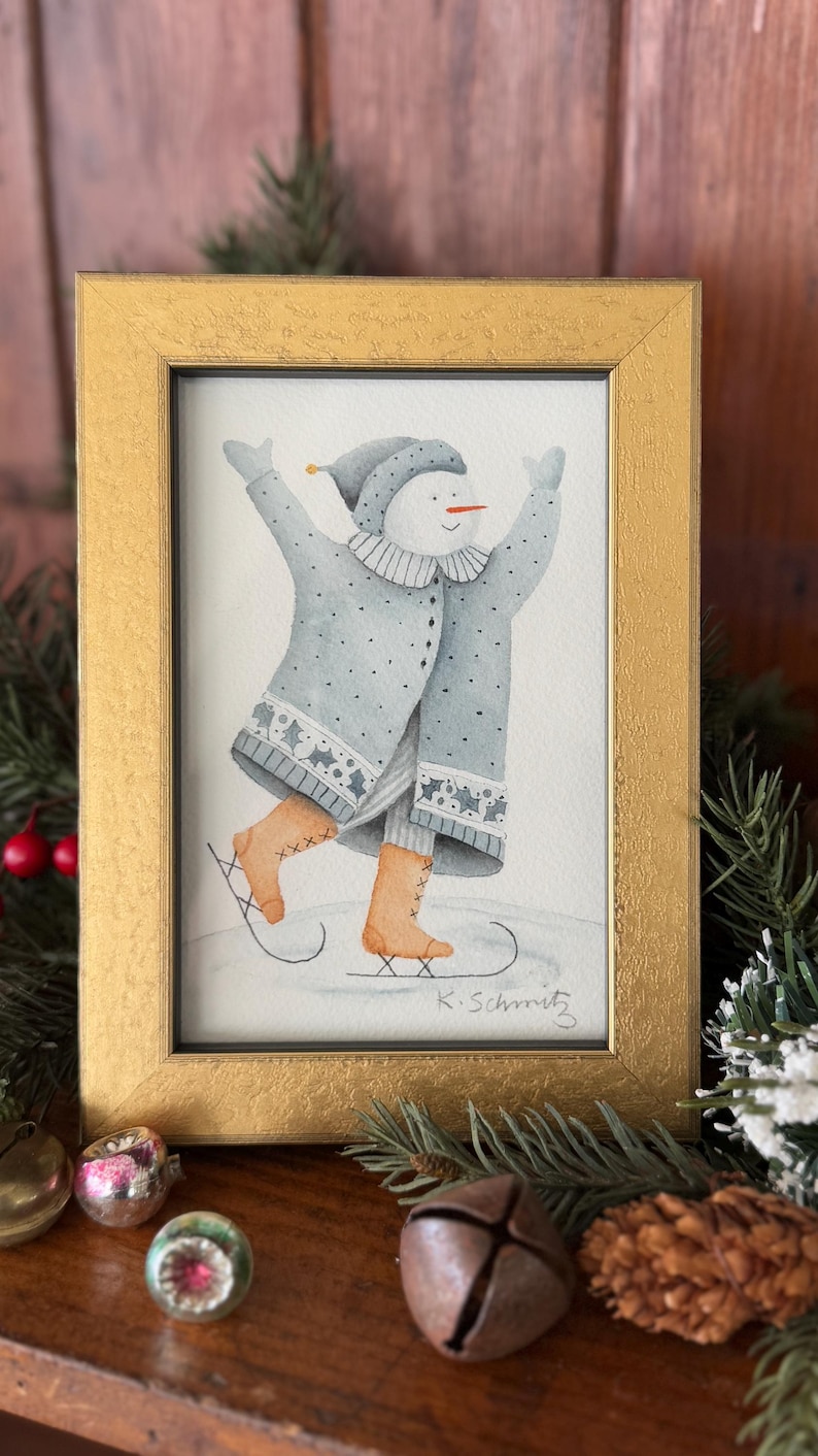 K&ouml;nnte beinhalten: Ein gerahmtes Aquarellgem&auml;lde eines Schneemanns beim Schlittschuhlaufen, der einen blauen Mantel und Hut sowie braune Schlittschuhe tr&auml;gt. Das Gem&auml;lde befindet sich in einem goldenen Rahmen, und die Signatur des K&uuml;nstlers befindet sich unten rechts. Weihnachtsdekorationen umgeben den Rahmen.