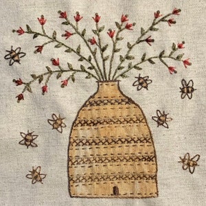 May include: Embroidered artwork featuring a tan vase filled with red flowers and green leaves. Surrounding the vase are several embroidered bees. The artwork is on a light beige fabric.