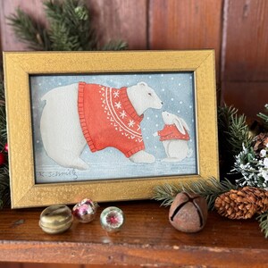 K&ouml;nnte beinhalten: Ein gerahmtes Aquarellgem&auml;lde mit einem Eisb&auml;ren und einem Hasen, die beide rote Pullover mit wei&szlig;en Schneeflockenmustern tragen. Das Gem&auml;lde ist vor einem schneebedeckten blauen Hintergrund und in einem goldfarbenen Rahmen gerahmt. Die Signatur des K&uuml;nstlers ist sichtbar.
