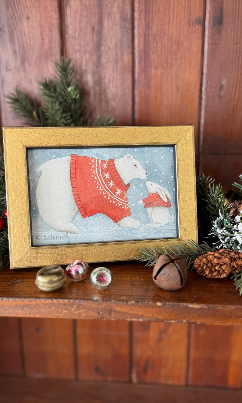K&ouml;nnte beinhalten: Ein gerahmtes Aquarellgem&auml;lde mit einem Eisb&auml;ren und einem Hasen, die beide rote Pullover mit wei&szlig;en Schneeflockenmustern tragen. Das Gem&auml;lde ist vor einem schneebedeckten blauen Hintergrund und in einem goldfarbenen Rahmen gerahmt. Die Signatur des K&uuml;nstlers ist sichtbar.