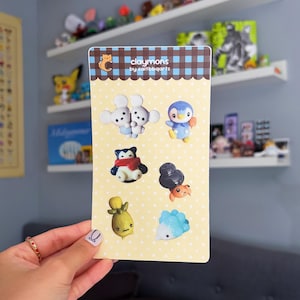 May include: A card featuring several small, colorful clay figurines. The card has a yellow background with white polka dots and the text "claymons by caitbbqarts". The figurines include mice, a penguin, and other creatures.