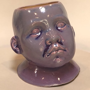 May include: A ceramic vase shaped like a baby's head, glazed in shades of purple. The vase has a wide opening at the top and is set on a small, rounded base. The face features detailed eyes, a nose, and a slightly open mouth.