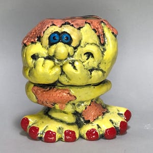 May include: A whimsical, hand-painted ceramic figurine in the shape of a troll. The troll has yellow skin, blue eyes, and red toenails. Its hair is a mix of pink and brown. The figure is in a seated position, with its hands clasped near its face.
