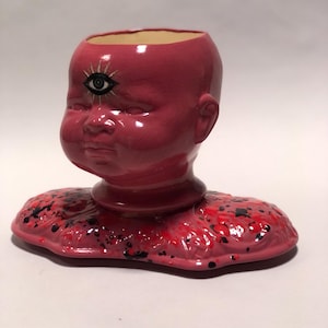 May include: A pink ceramic sculpture of a baby's head with an open top and a third eye. The sculpture has a base with red and black splatters. The eye is black with gold accents. The sculpture is a unique piece of art.