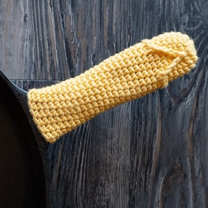 Puede incluir: Una funda de mango de ganchillo amarilla para una sartén de hierro fundido. La funda está hecha con un punto sencillo y es una excelente manera de proteger tus manos del calor.