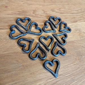 May include: A collection of small, handcrafted metal hearts arranged on a wooden surface. The hearts are dark grey with a textured finish, and they are arranged in a heart shape. The background is a light brown wooden surface.
