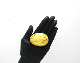 May include: A yellow, speckled egg-shaped object with red spots, held in a black gloved hand against a white background. The egg is smooth and oval, with a glossy finish. The overall theme is decorative.