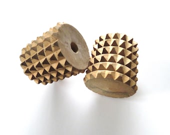 May include: Two wooden massage rollers with a geometric, pyramid-like design. The rollers are light brown and appear to be made of wood. One roller is lying on its side, while the other stands upright.