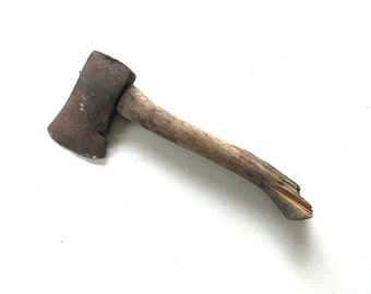 May include: A small, vintage axe with a weathered wooden handle and a rusty, metal head. The axe is positioned diagonally against a white background, showcasing its aged appearance and design. The handle is approximately 12 inches long.