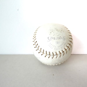 Puede incluir: Una pelota de softball de cuero blanco con costuras marrones y el logotipo de Spalding y "751" impreso en ella.