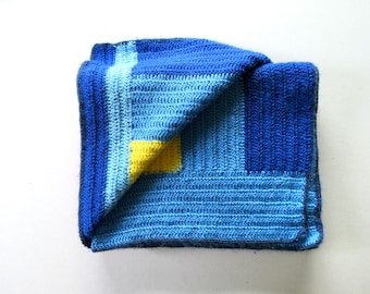 May include: A folded, handmade crochet blanket in shades of blue and a small yellow square. The blanket features a geometric design with varying textures and tones of blue, creating a cozy and visually appealing textile.
