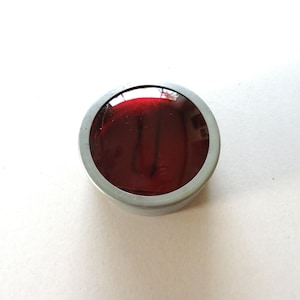 May include: A round, red glass object with a silver-colored metal rim. The glass has a reflective surface, and the object is set against a white background. The item appears to be a decorative piece or component.