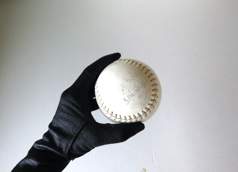 Puede incluir: Una pelota de softball blanca con la palabra "SPALDING" escrita en negro. La pelota se sostiene en un guante negro.