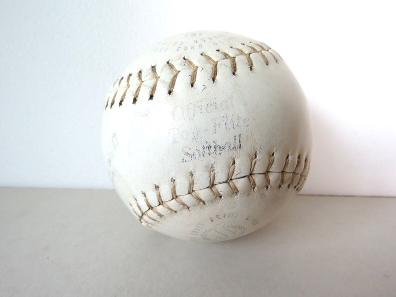 Puede incluir: Una pelota de softbol de cuero blanco con costuras marrones. La pelota tiene el texto "Official Softball" y "12" impreso en ella.