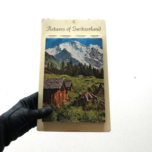 May include: Vintage postcard featuring a scenic view of a snow-capped mountain range with a small village in the foreground. The postcard is titled "Pictures of Switzerland".