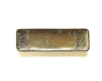 May include: A rectangular, antique brass tray with ornate, symmetrical designs at each end. The tray has a slightly tarnished appearance, suggesting age and use. The decorative elements feature swirling patterns and stylized floral motifs.