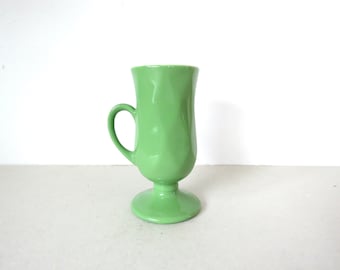 May include: A light green ceramic mug with a handle. The mug has a pedestal base and a tall, slightly flared body. The mug is approximately 6 inches tall and is a solid, glossy color.