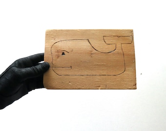 May include: A wooden puzzle piece in the shape of a whale. The whale outline is etched into a rectangular piece of light-colored wood. The puzzle is held by a gloved hand against a white background.
