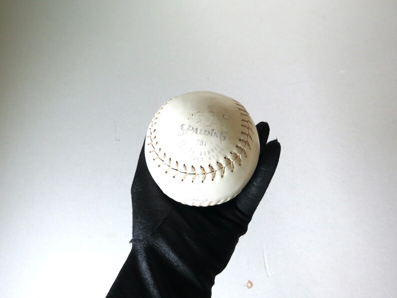 Puede incluir: Una pelota de softball blanca con costuras marrones y el logotipo de Spalding y "751" impreso en ella.