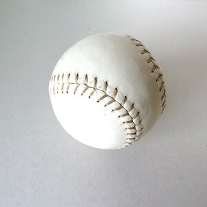 Puede incluir: Una pelota de softbol de cuero blanco con costuras marrones.