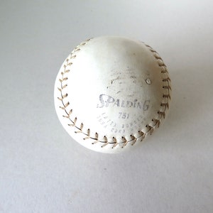 Puede incluir: Una pelota de softbol de cuero blanco con costuras marrones y el logotipo de Spalding y "751" impreso en ella.