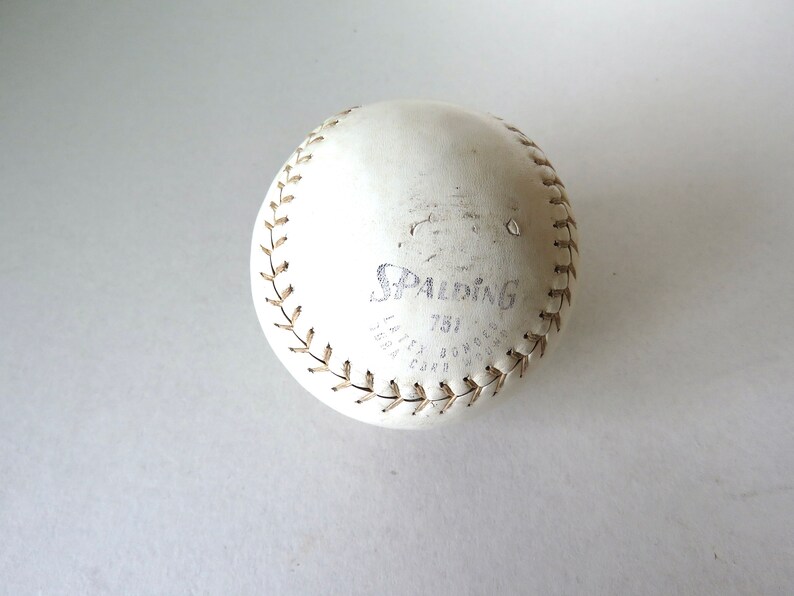 Puede incluir: Una pelota de softbol de cuero blanco con costuras marrones y el logotipo de Spalding y "751" impreso en ella.