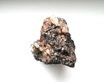 May include: A rough, natural mineral specimen with a mix of colors. The rock features dark gray, black, and brown tones, with crystalline formations and lighter, beige-colored areas. The overall texture appears coarse and uneven.