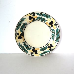 May include: A white ceramic plate with a green and yellow floral design. The flowers are black with yellow centers and are surrounded by green leaves.