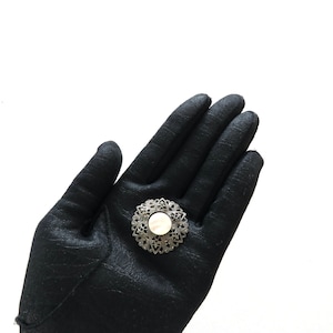 May include: A silver-toned brooch with an intricate openwork design surrounding a central, round, mother-of-pearl-like insert. The brooch is held against a black background, highlighting its delicate details and vintage aesthetic.