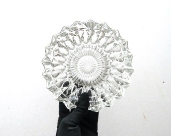 May include: Clear glass ashtray with a starburst design in the center and a geometric, faceted rim. The ashtray is round and held by a gloved hand against a white background.