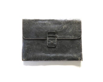 May include: A distressed black leather clutch with a flap closure and a rectangular buckle. The clutch has white stitching around the edges and a worn appearance. The bag is likely vintage or has a vintage aesthetic.