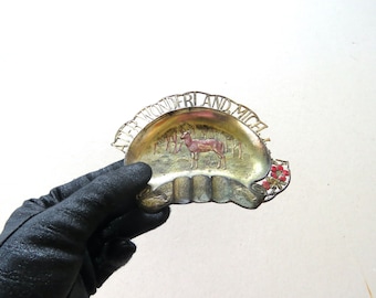 May include: Vintage metal ashtray with a deer and forest scene. The ashtray is shaped like a half-moon and has the words "WONDERLAND MICH" in a decorative font. Red and pink floral accents are on the right side.