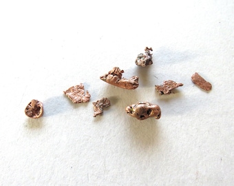 May include: Close-up of several small, irregularly shaped pieces of copper metal on a white surface.