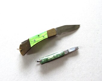 May include: Two pocket knives are displayed against a white background. The larger knife has a green and brass handle, and the blade is open. The smaller knife has a green handle and a closed blade. Both knives have a small ring for attaching a lanyard.