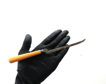 May include: A vintage fish knife with a serrated blade and a light brown handle. The knife is held against a black glove, showcasing its design and craftsmanship. The blade is curved, and the handle is smooth.