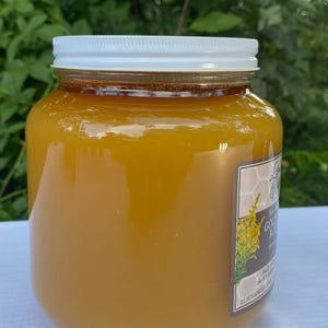 May include: A jar of golden-colored honey with a white lid. The jar has a label with text and a floral illustration. The honey is thick and appears to be freshly harvested. The jar is sitting on a white surface.