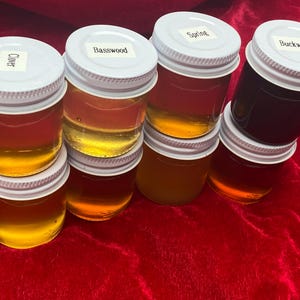 May include: Eight small glass jars with white lids filled with different types of honey. The jars are labeled with the type of honey: Clover, Basswood, Spring, Buckwheat.