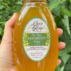 May include: A glass jar of raw basswood honey from Locust Ridge Apiaries. The jar is labeled with the company name, product name, weight, and contact information. The honey is a light amber color.